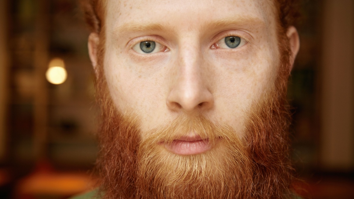 Highly-detailed portrait of good-looking Caucasian hipster with long red beard, beautiful blue eyes and freckles standing against blurred background of cozy cafeteria.