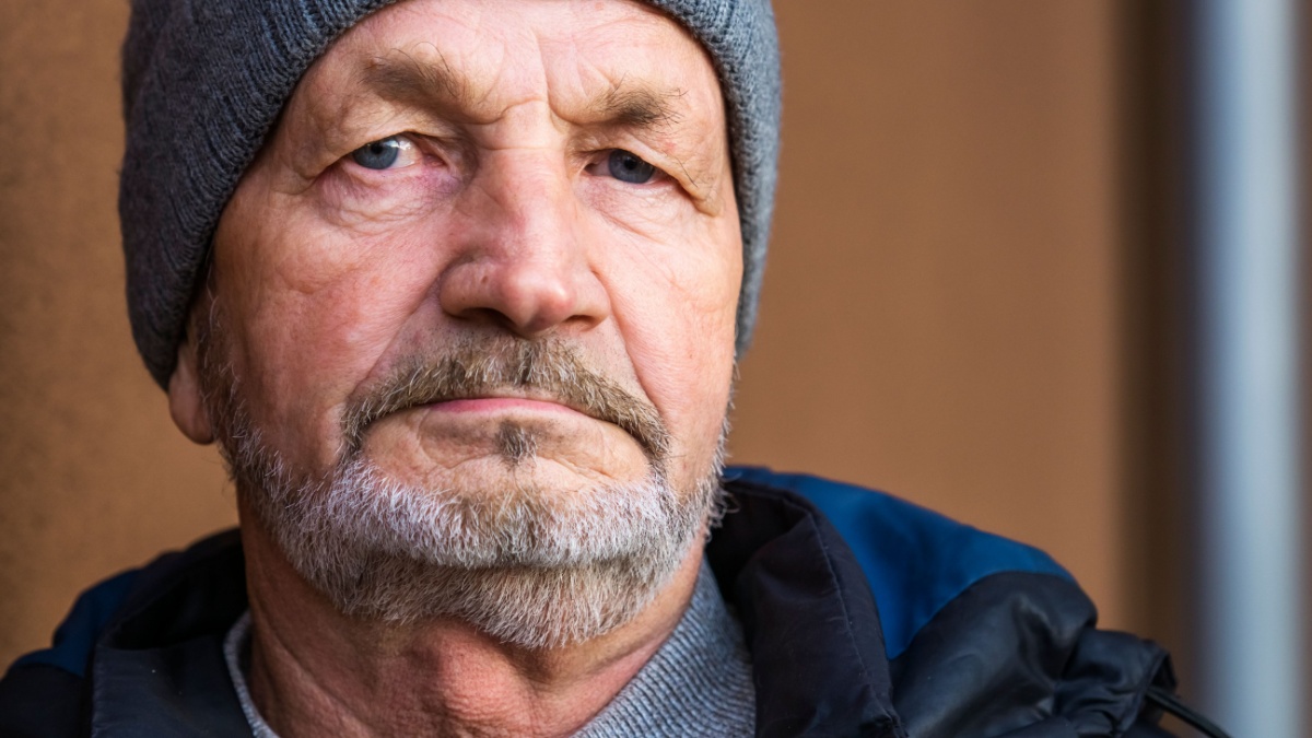 An elderly bearded pensive man in an autumn jacket and knitted hat on the background wall of an apartment building.