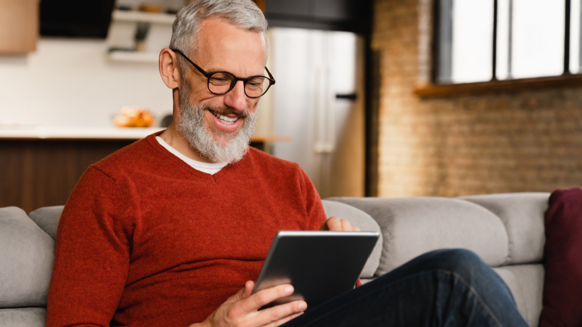 Smiling caucasian middle-aged mature man in glasses using digital tablet for e-learning, paying bills online, e-commerce, remote work, social media online at home.