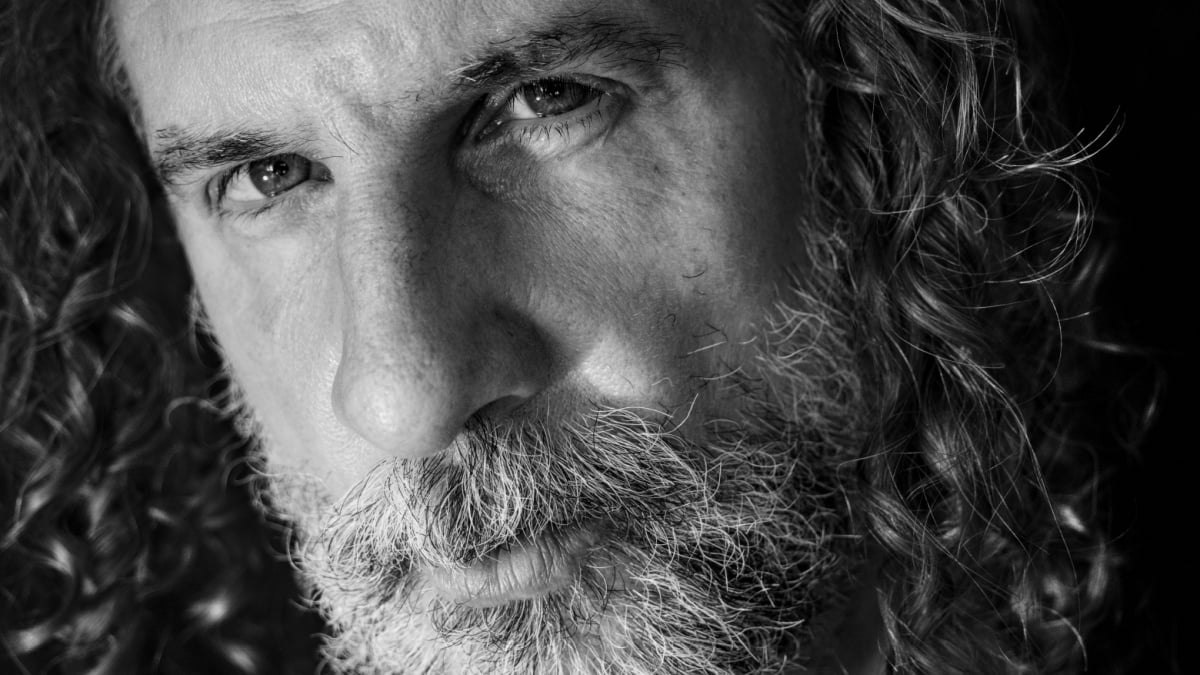 Black and white close up portrait of a 45 years old man, with long curly hair, salt and pepper beard, on a black background..