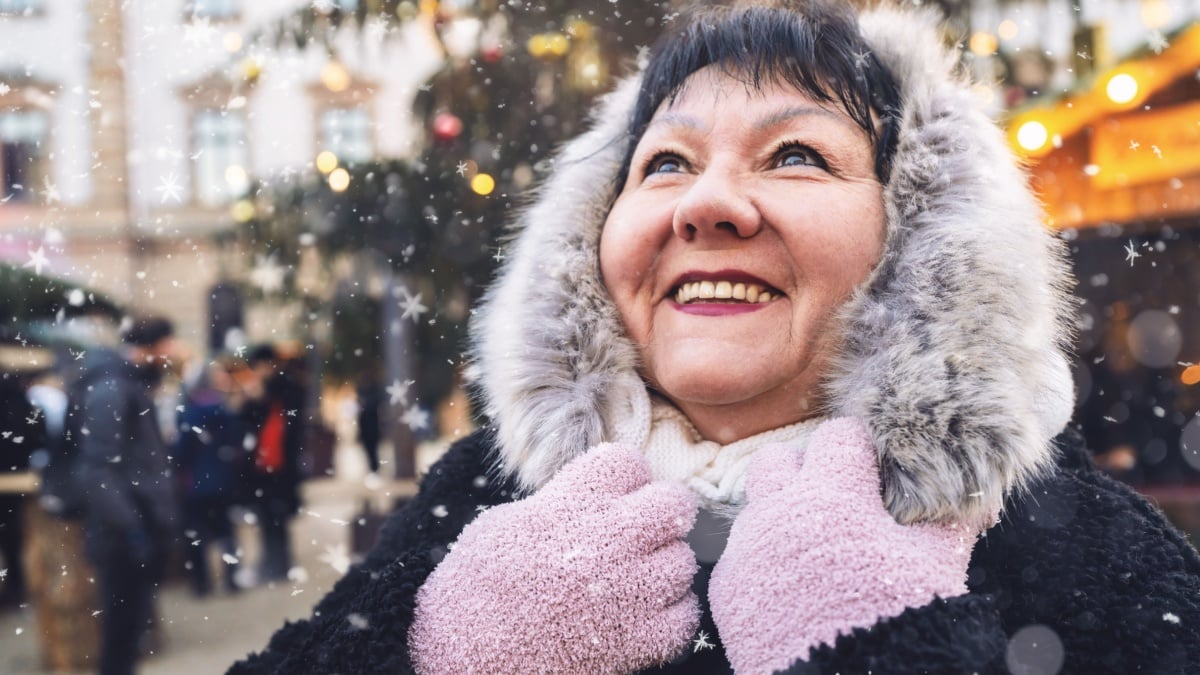 Elderly woman smiling at a winter Christmas market, with snow falling, in festive attire.