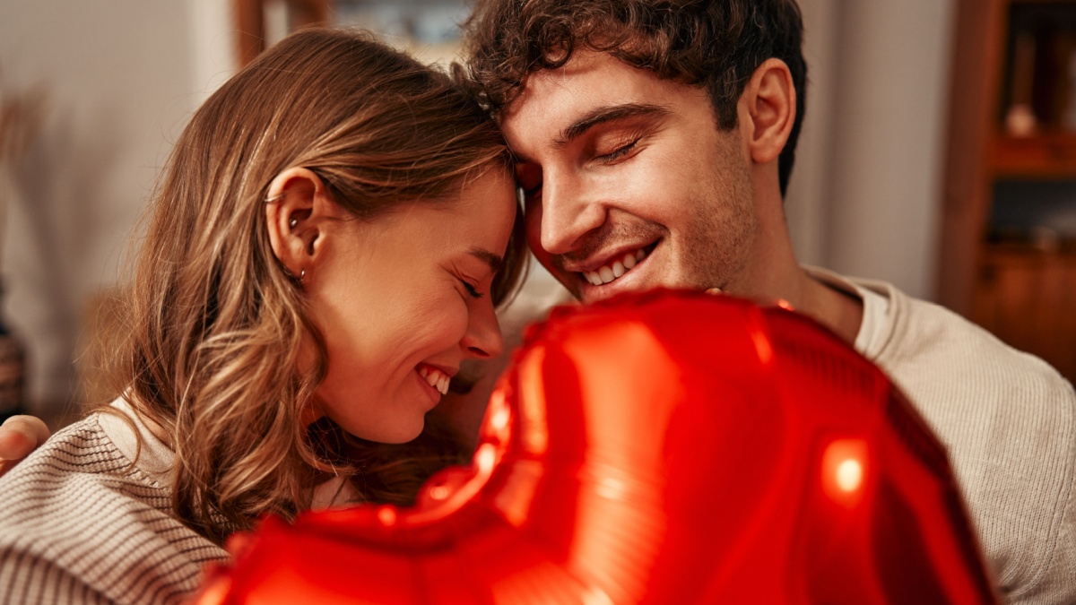 Happy Valentine's Day. Young couple in love holding a heart-shaped balloon while sitting on the sofa in the living room at home. Romantic evening together.
