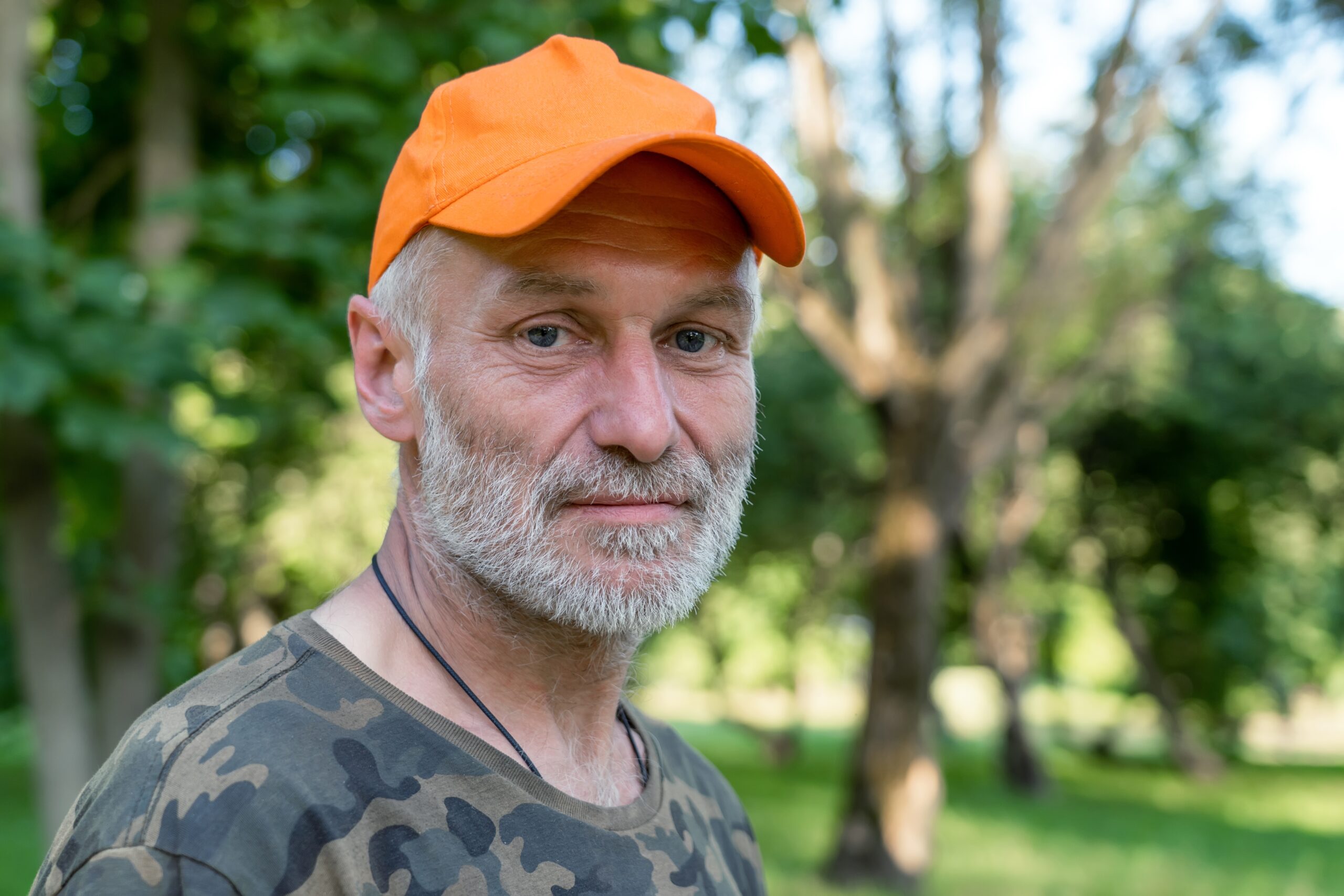 Close up portrait of sad mature man wearing orange cap looking at camera, standing in park outdoors. Senior caucasian man with grey beard.