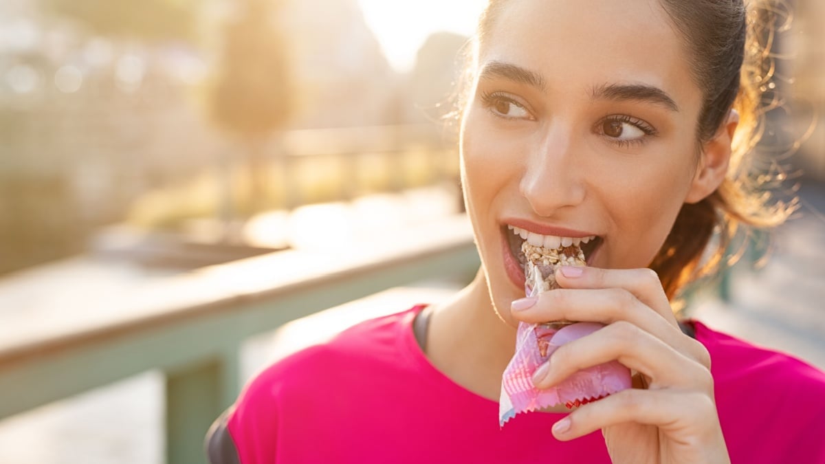 Athletic woman eating a protein bar. Closeup face of young sporty woman resting while biting a nutritive bar. Fitness beautiful woman eating a energy snack outdoor.