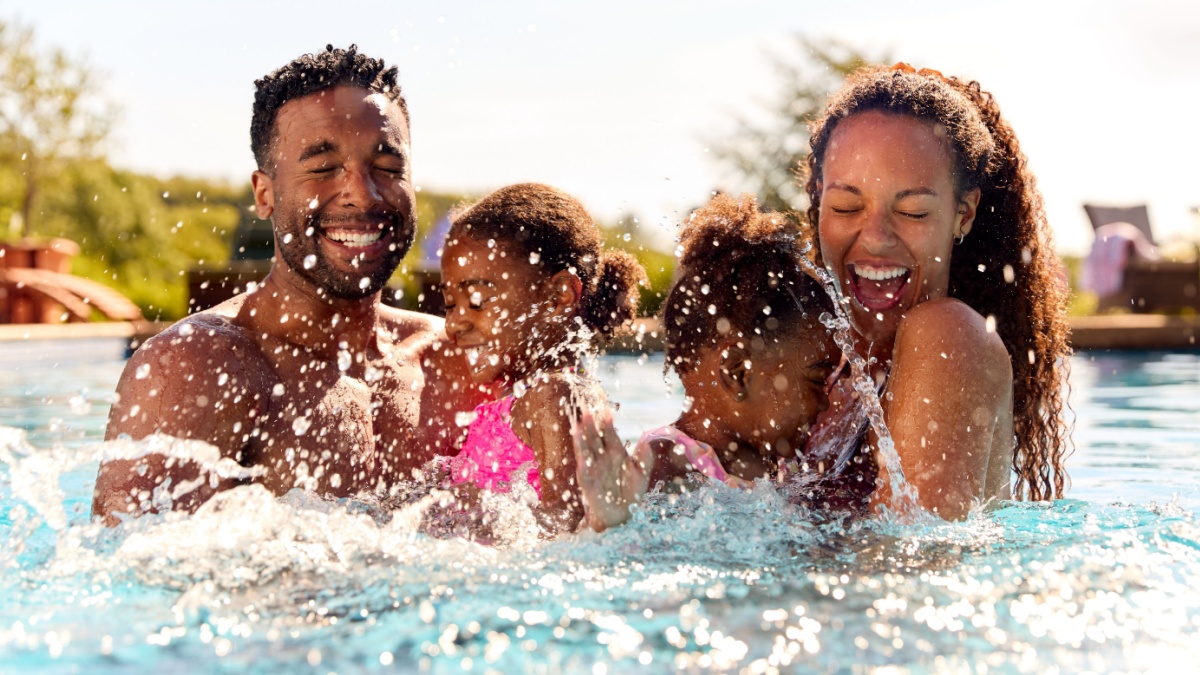 Family On Summer Holiday With Two Girls Being Held In Swimming Pool By Parents And Splashing.