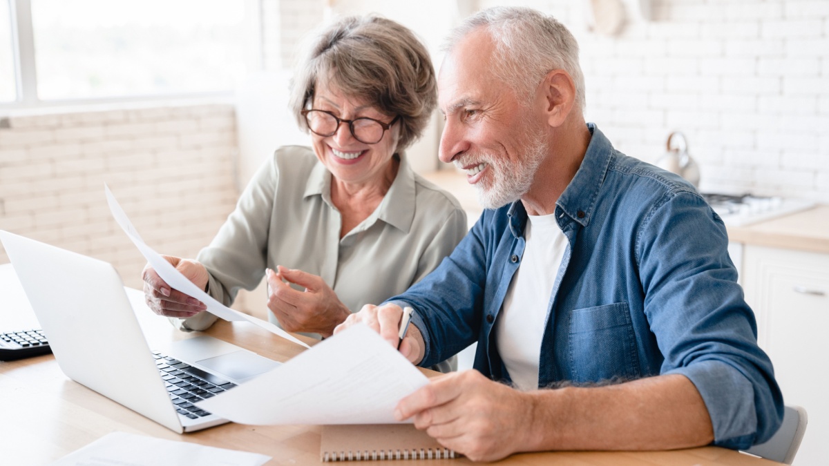 Smiling caucasian old elderly senior couple husband and wife using laptop and doing paperwork together at home kitchen. E-banking, e-commerce.