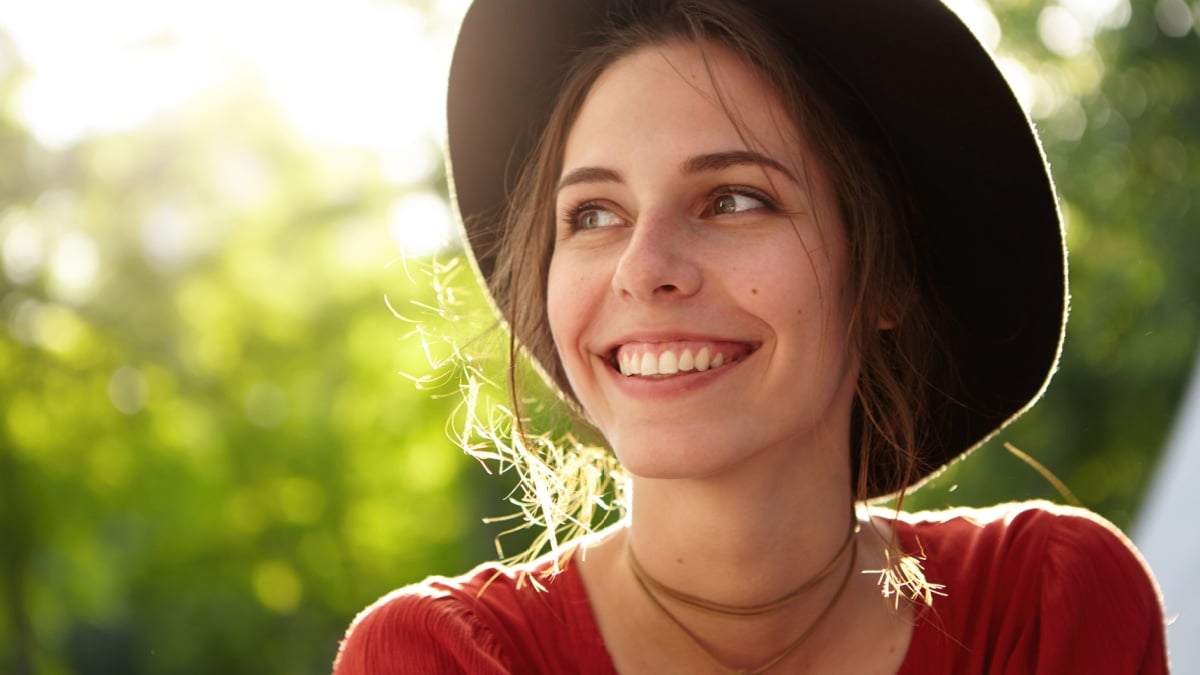 People, nature, resort, summer, vacations concept. Sunny portrait of cute young female in trendy hat smiling pleasantly looking aside taking pleasure from bright sunlight and gentle summer wind.