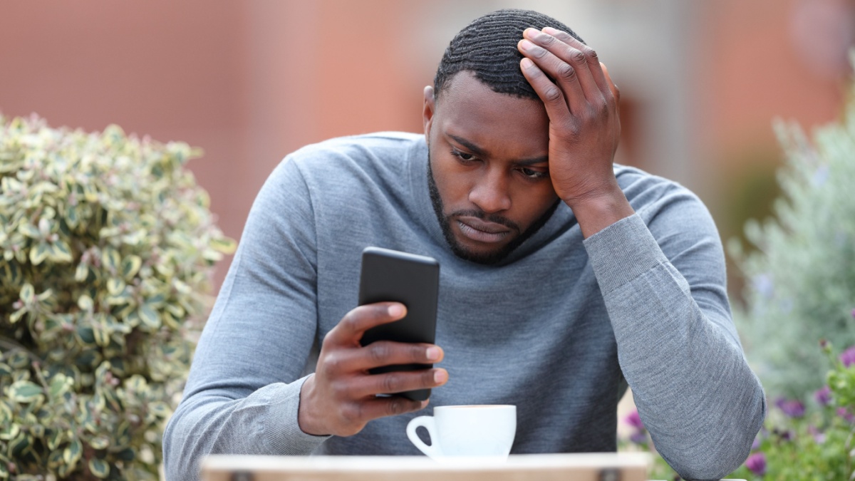 Front view portrait of a worried man with black skin checking phone in a coffee shop.