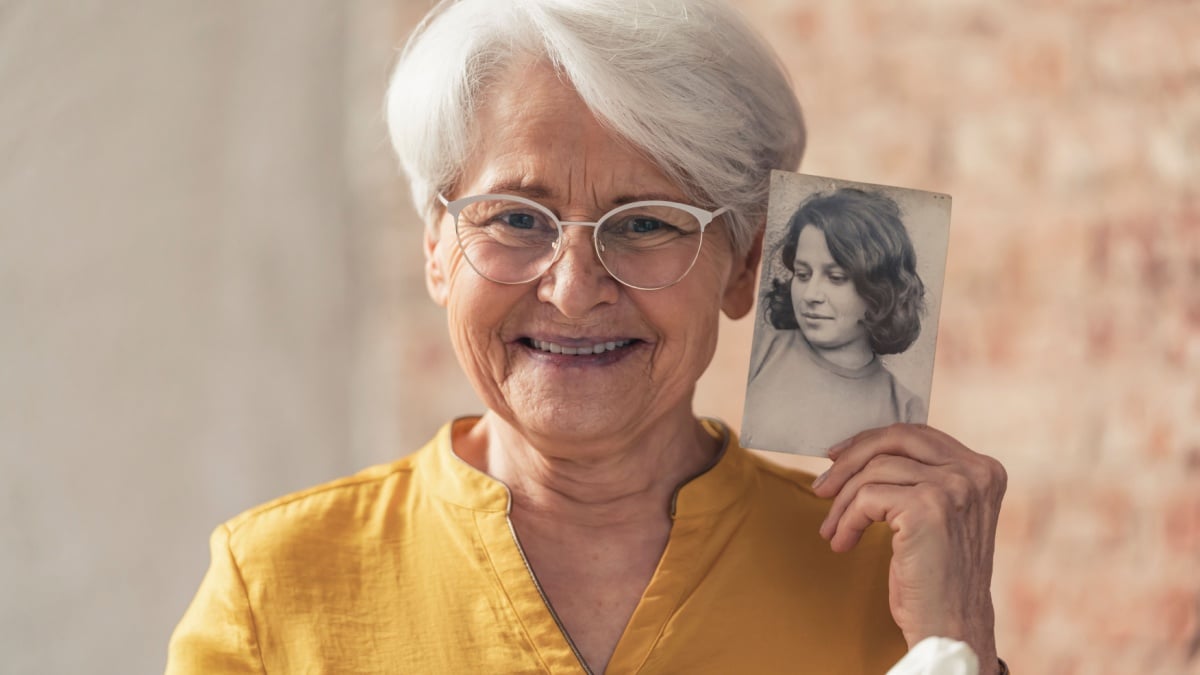 smiling elderly lady with short grey hair comparing her current face to a photo of her younger self.