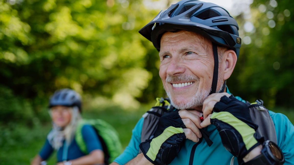 Active senior couple riding bicycles at summer park, man is putting on helmet, healthy lifestyle concept.
