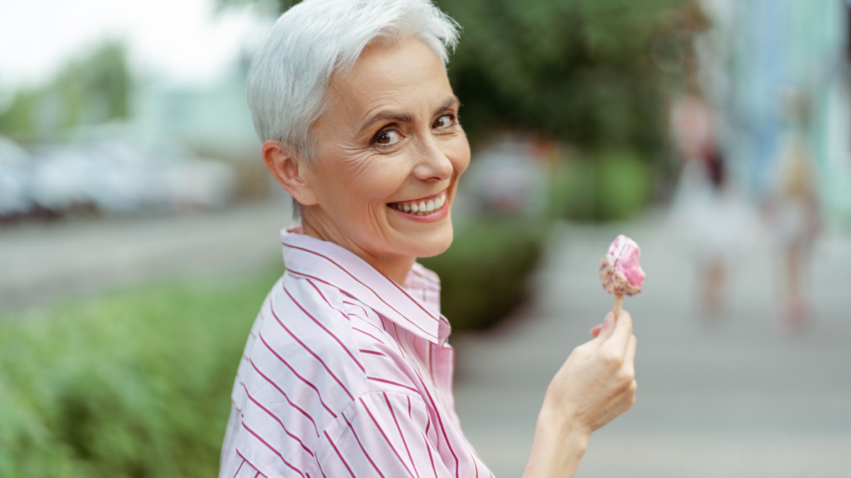 Cheerful stylish gray haired woman looking at camera while eating tasty ice cream in park. Positive lifestyle and tasty food concept.