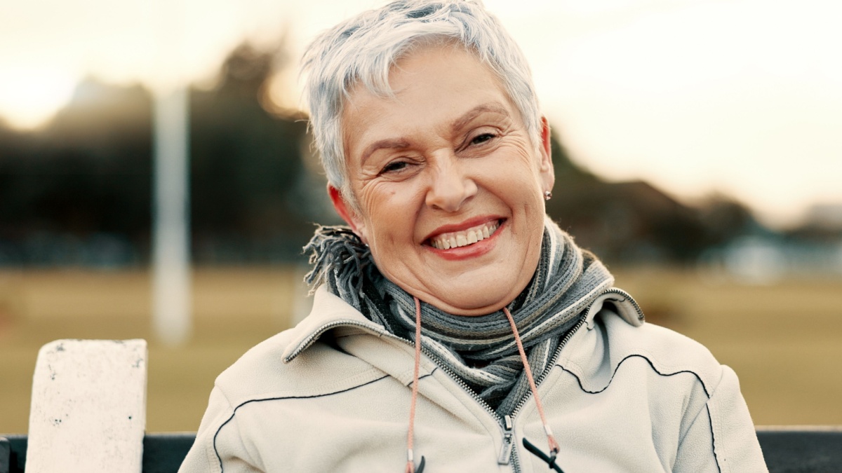 Face, senior woman and funny on park bench on vacation, holiday or travel in winter. Portrait, happy and elderly person in nature, outdoor or garden for laughing, freedom and retirement in Australia.