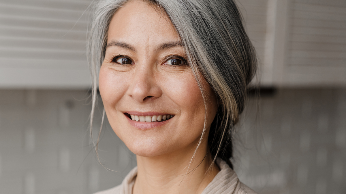 Mature grey woman smiling while drinking water in kitchen at home.