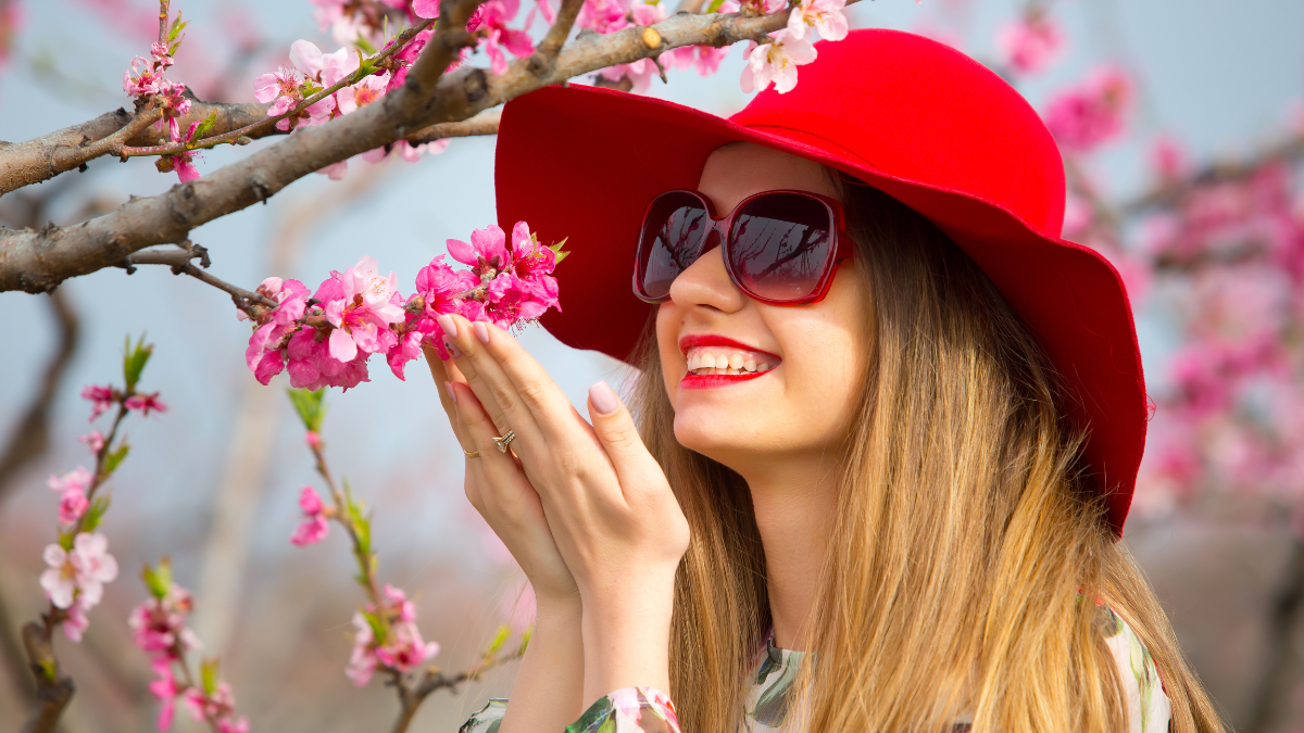 Smiling face of a beautiful girl with floral dress and big red hat standing on green grass in a garden with blooming trees. Spring time.