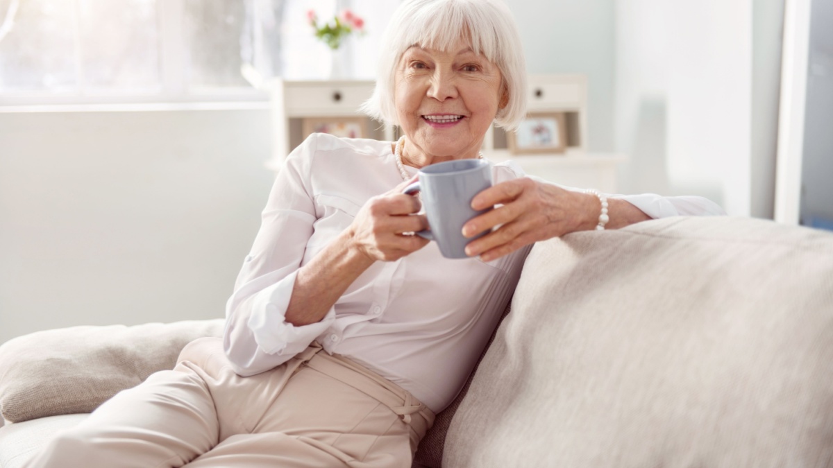 Delicious drink. Cheerful elderly woman sitting on the sofa in her living room and holding a cup of coffee while smiling at the camera.