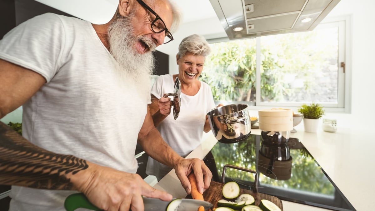 Happy senior couple having fun cooking together at home - Elderly people preparing health lunch in modern kitchen - Retired lifestyle family time and food nutrition concept.