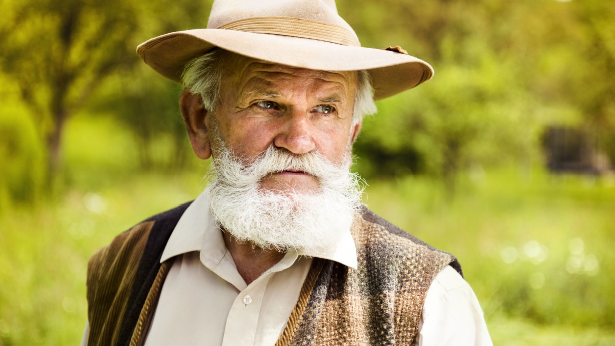 Portrait of old farmer with beard and hat in his backyard.