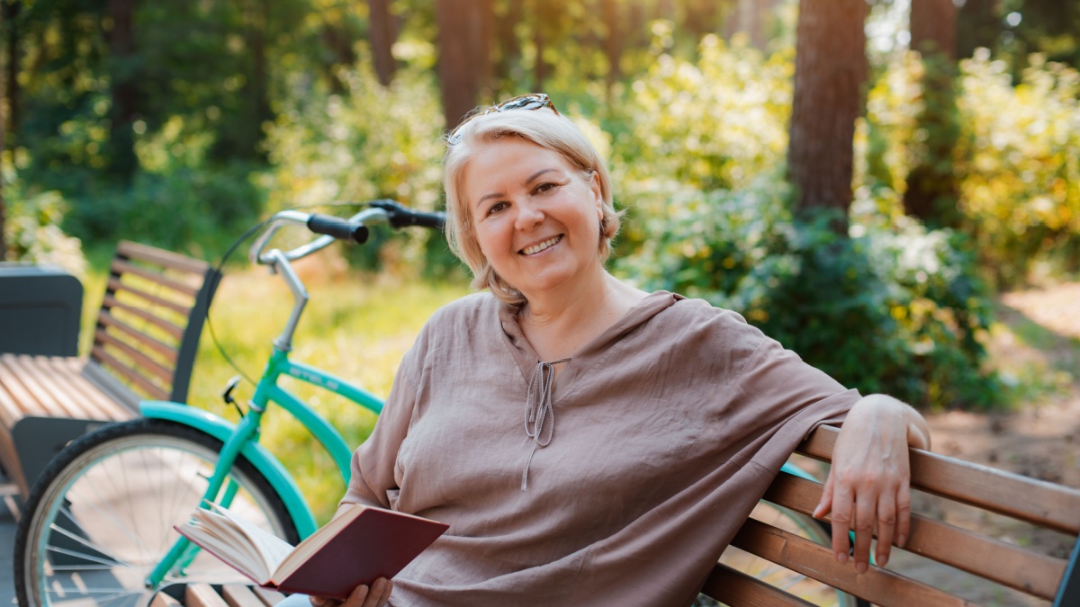 smiling modern old lady relaxing city park. Pensive senior gray haired woman casual sitting bench outdoors reading book. cycling forest park, bicycle, healthy active lifestyle after 50-60 years.