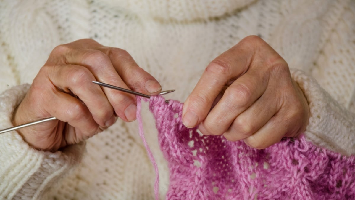 grandmothers hands knit pink wool yarn. craft is hobby of old women. senior lady, granny in white knitted sweater handmade. needle pattern from thread loop. close-up warm, traditional, domestic textil.