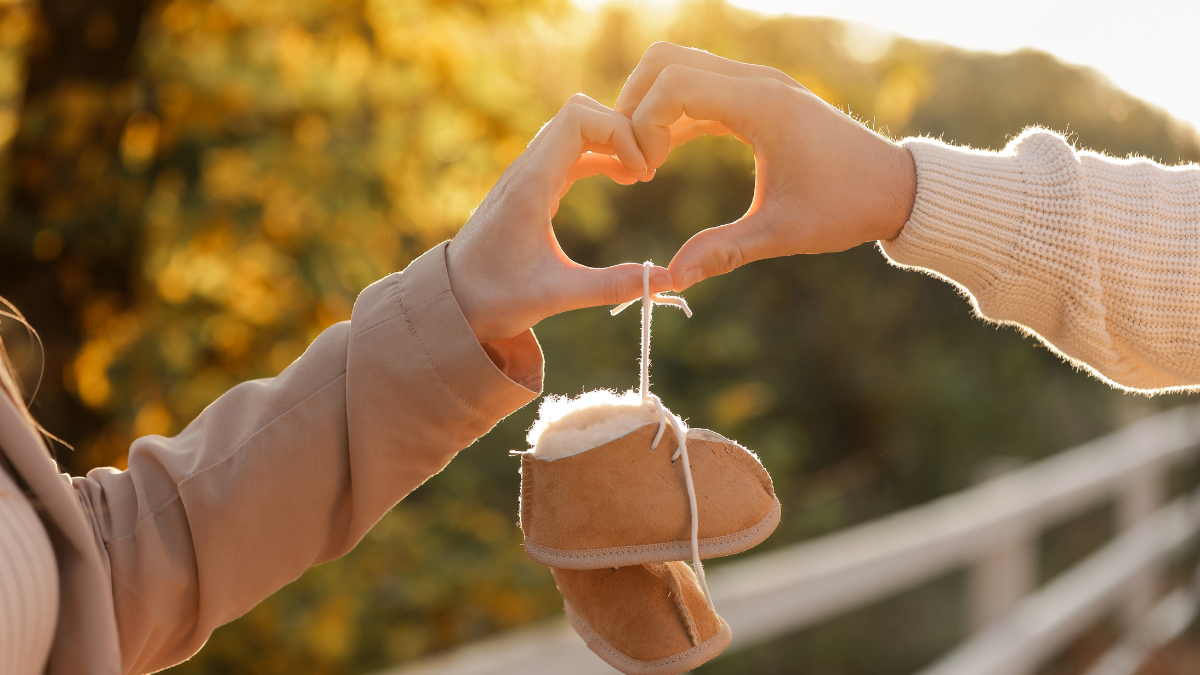 Man and pregnant wife are doing heart gesture with hands and holding warm baby shoes on nature autumn background. Future parents waiting for the baby.