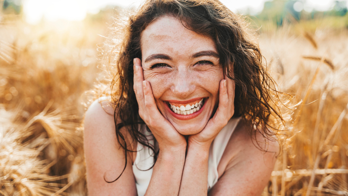 Happy beautiful woman smiling at camera in a wheat field - Delightful female enjoying summertime sunny day outside - Wellbeing, mental health, body care and happiness concept.