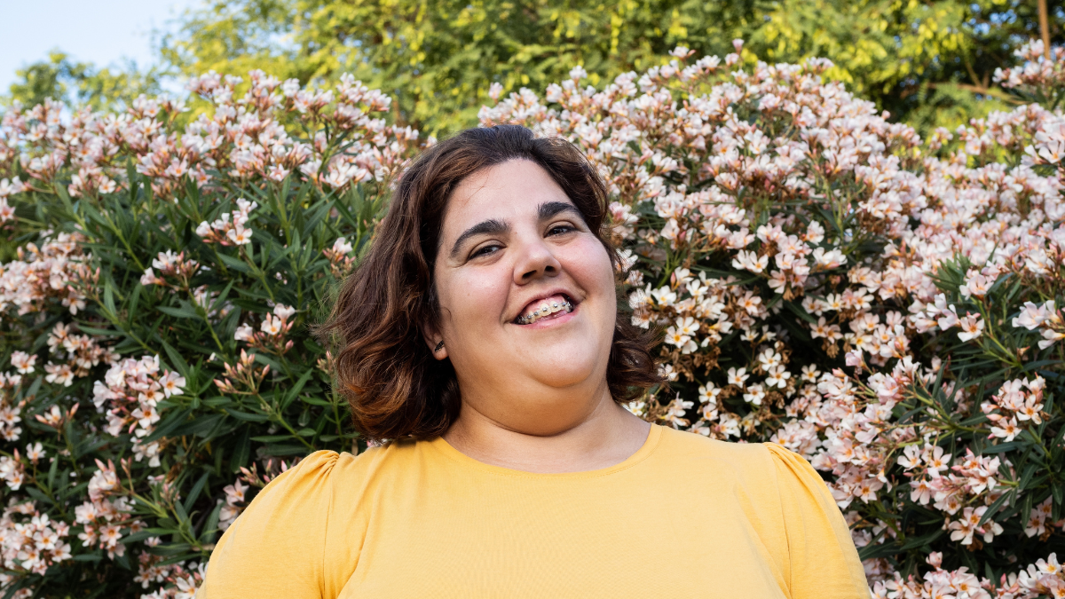 A joyful woman with neurodiversity smiles in front of flowering bushes in a garden during the afternoon.