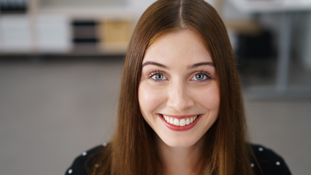 Gorgeous vivacious young woman with a beaming smile and happy eyes smiling directly at the camera, close up head and shoulders portrait indoors.