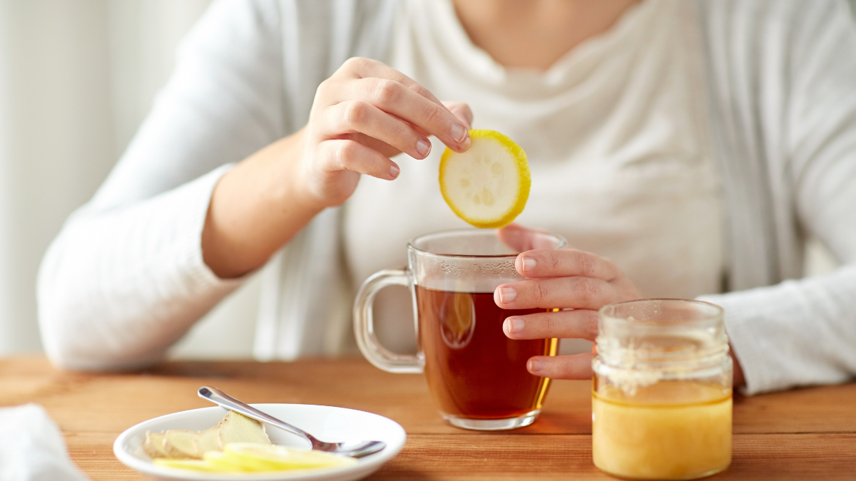 health, traditional medicine and ethnoscience concept - close up of ill woman drinking tea with lemon, honey and ginger at wooden table.