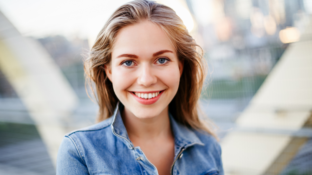 Closeup portrait of beautiful smiling blonde white Caucasian girl woman with long hair wearing jeans jacket outside in evening night city street bridge looking in camera.