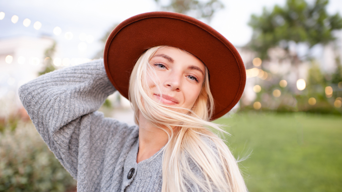Smiling young beautiful blonde woman wear hat and knitted woolen jumper posing over city street in park outdoors close up. Happy pretty teenage girl 18-19 year old having fun outside. 20s. Happiness.