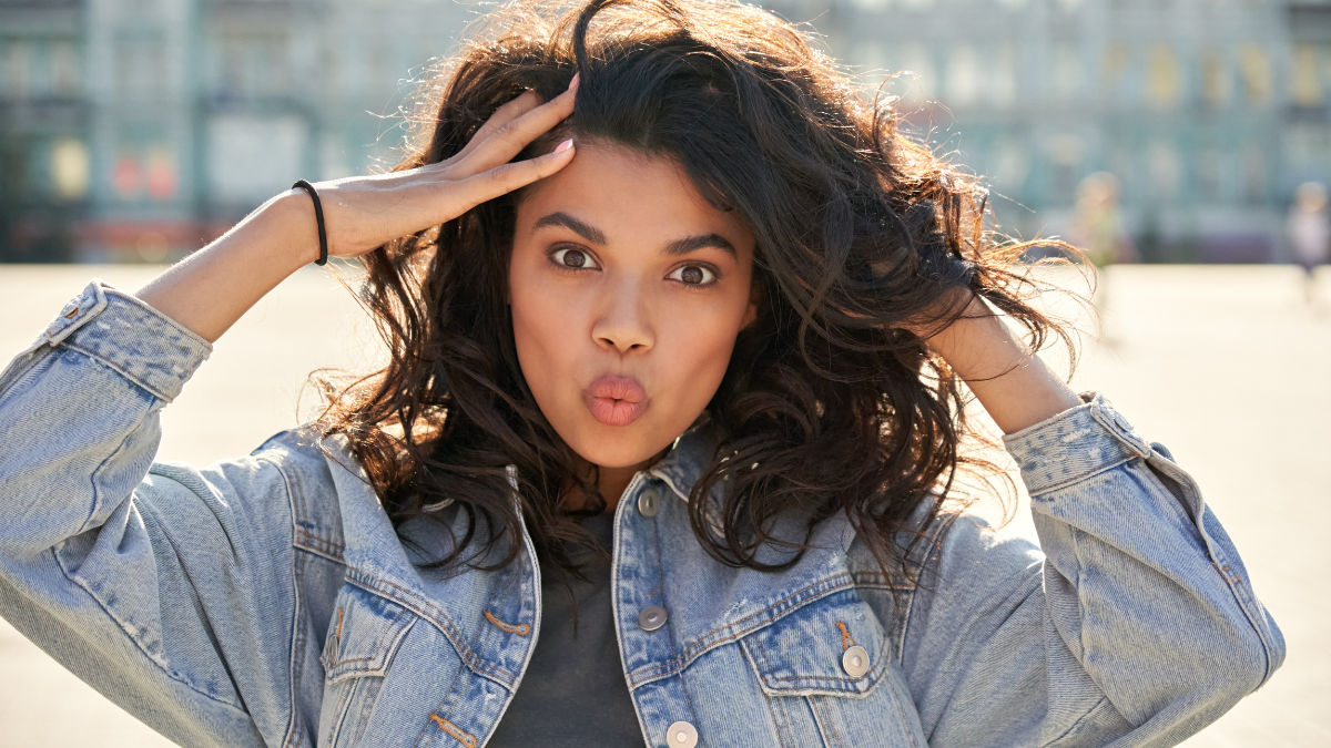 Pretty cool African American young woman wears denim jacket pouting lips stands on sunny city street backlit with sunlight. Curly funny young black lady looks at camera outdoor, urban portrait.