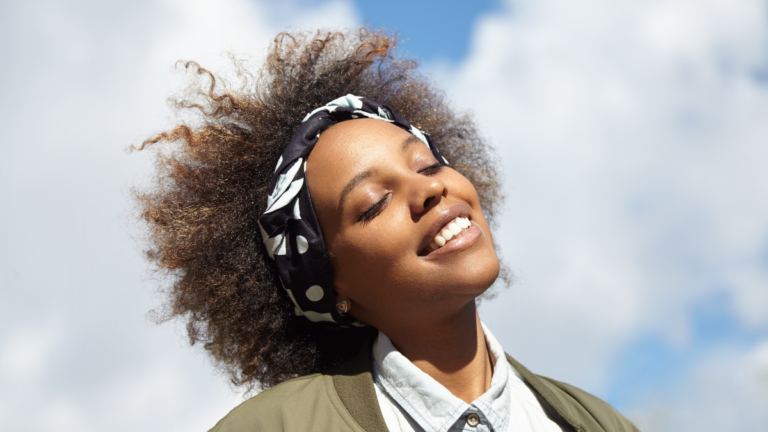 Close up shot of young black hipster student wearing do-rag on her head, smiling and closing her eyes with happy face expression, dreaming and enjoying warm summer wind while walking outdoors alone.