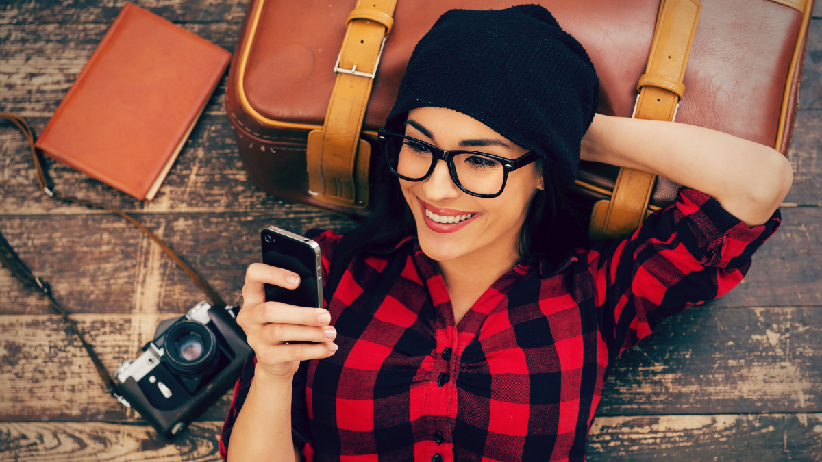 Relaxing after long trip. Top view of beautiful young woman in headwear lying on the floor holding mobile phone and smiling.