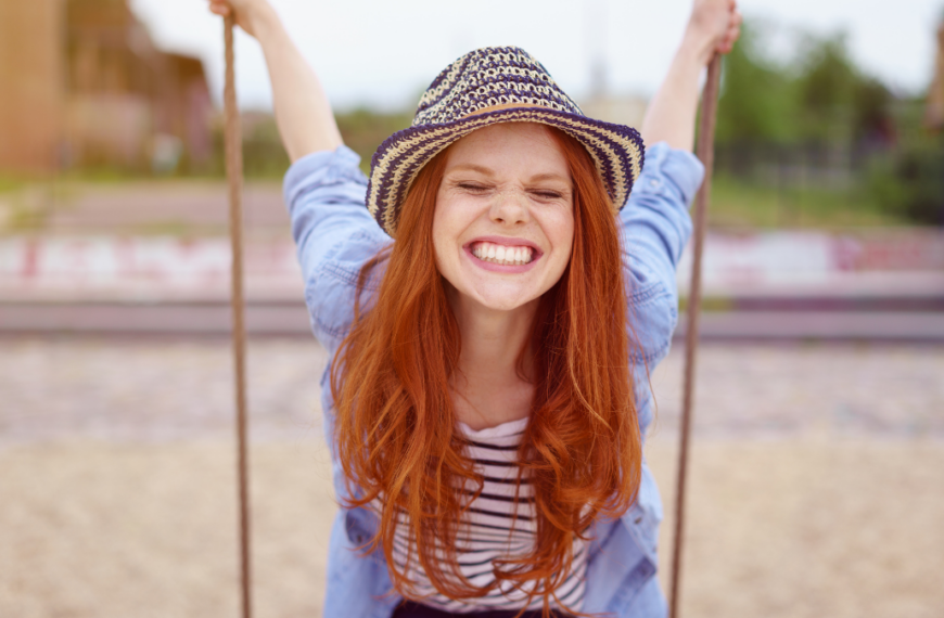 Overjoyed single red haired young woman in hat and blue denim shirt with rolled up sleeves sitting on swing at urban playground.