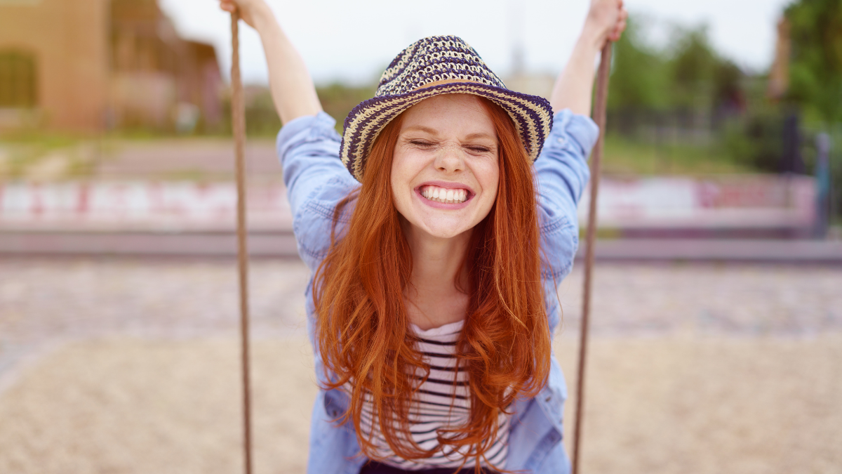 Overjoyed single red haired young woman in hat and blue denim shirt with rolled up sleeves sitting on swing at urban playground.