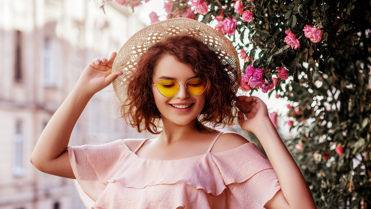 Outdoor close up portrait of young beautiful happy smiling curly girl wearing stylish yellow sunglasses, straw hat, pink blouse with ruffles. Model posing near blooming roses. Summer fashion concept.