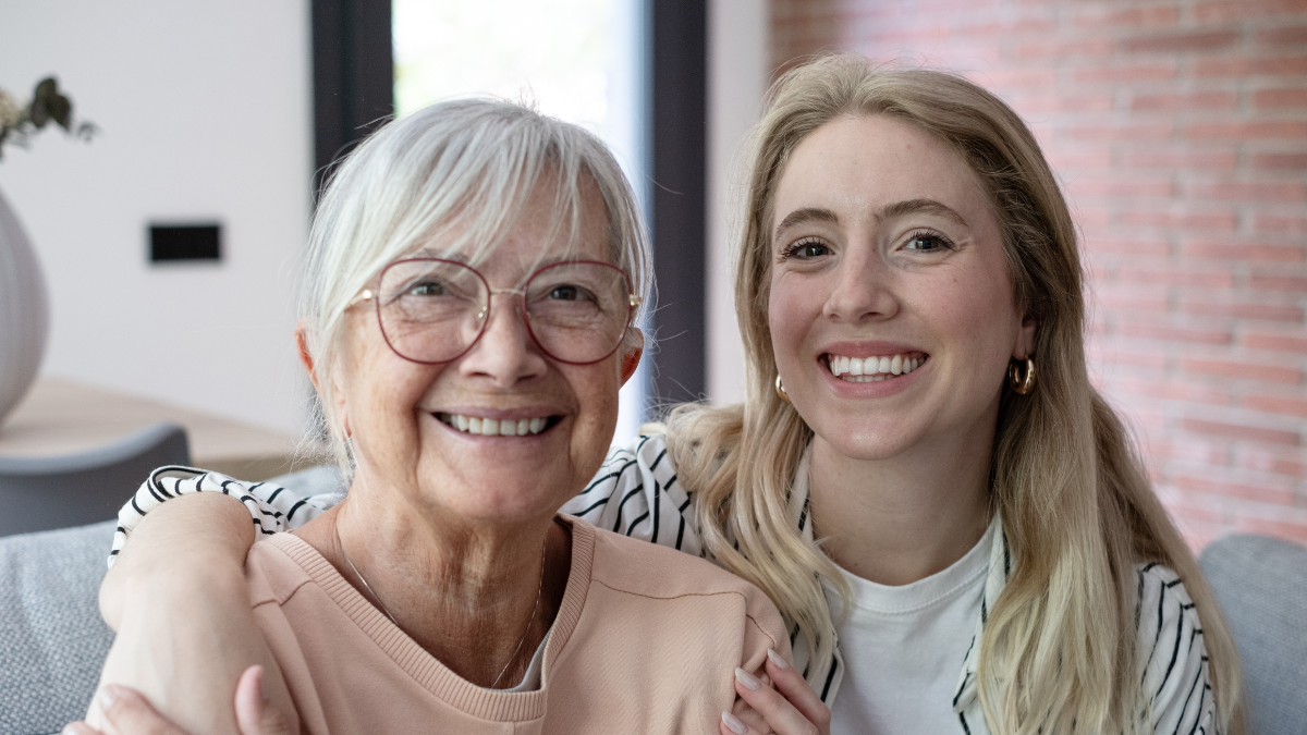 A happy grandmother and granddaughter sit closely on a living room sofa, smiling and embracing warmly, conveying family love, multigenerational bond, affection, comfort and joyful companionship at home. Old-Fashioned Frugal Living Tips Your Grandparents Used to Stretch Every Dollar.