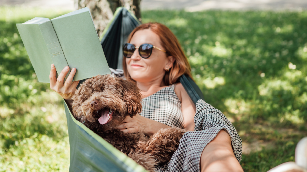 Woman reading book relaxing in hammock with her fluffy brown Maltipoo dog on sunny day. Both looking content and happy. This outdoor scene captures joy of bonding with pets and enjoying togetherness.
