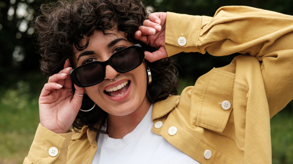 Fashionable young woman with curly hair, nose piercing, and large hoop earrings smiles while adjusting sunglasses and wearing a stylish yellow jacket outdoors.