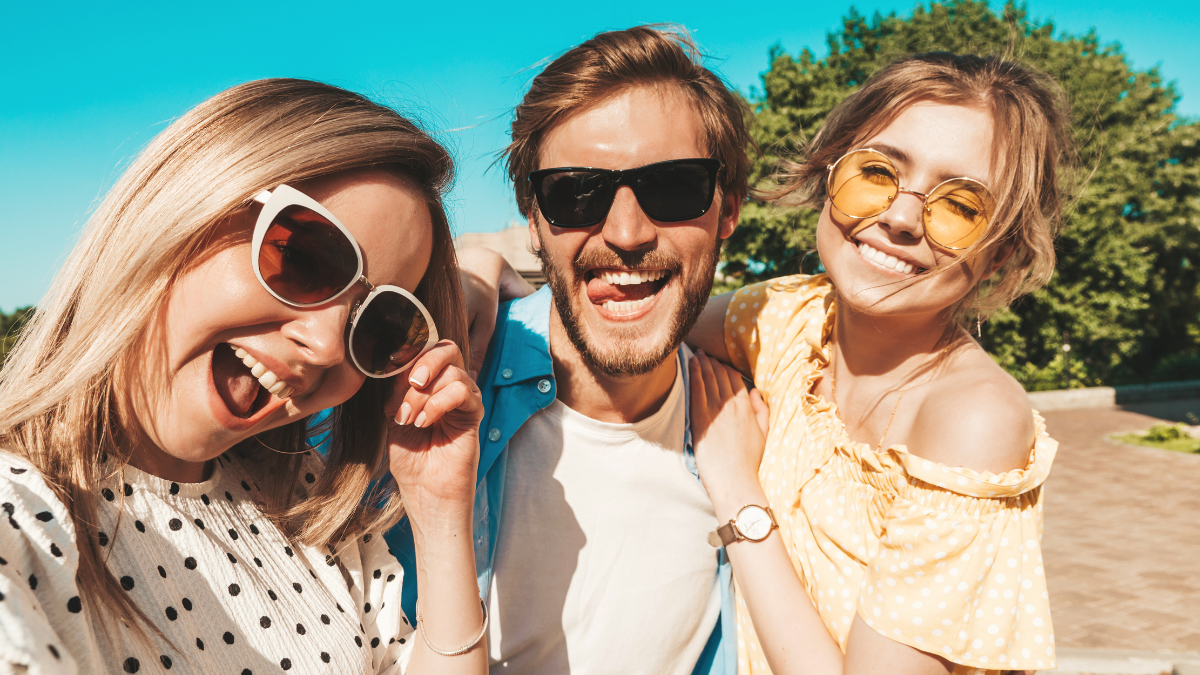 Group of young three stylish friends in the street.Man and two cute girls dressed in casual summer clothes.Smiling models having fun in sunglasses.Women and guy making photo Pov selfie on smartphone.
