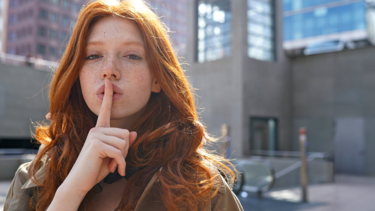 Hipster teen stylish cool redhead fashion girl model showing shh sign asking to keep secret, be hush silent or privacy silence standing in big city urban location. Headshot portrait.