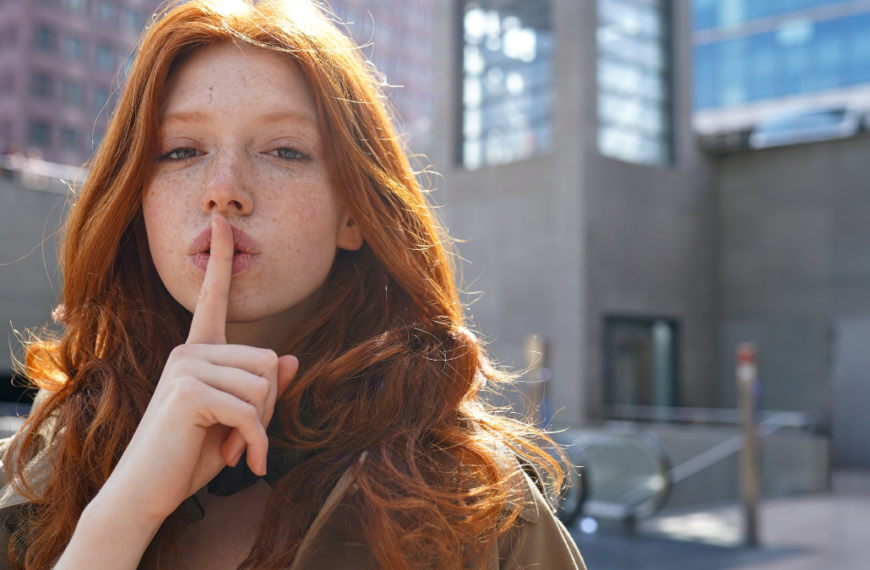 Hipster teen stylish cool redhead fashion girl model showing shh sign asking to keep secret, be hush silent or privacy silence standing in big city urban location. Headshot portrait.