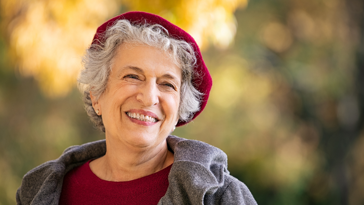 Portrait of senior woman smiling outdoor. Cheerful old woman in autumn park with copy space. Happy retired lady with grey hair and red beret smiling and looking away.