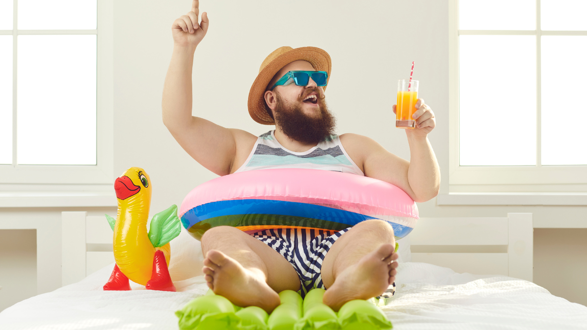 Funny guy in sunglasses, sun hat and swim ring sitting on beach mattress and sipping cocktail points finger up struck by cool idea on how to turn quarantine holiday at home into fun summer vacation.