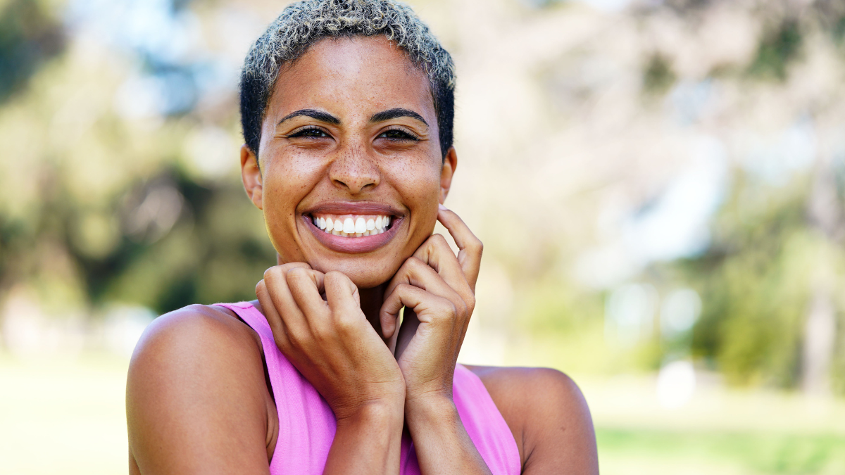 Beautiful young multicultural woman with short hair and hands up to face is standing at the park and smiling because she's super happy.
