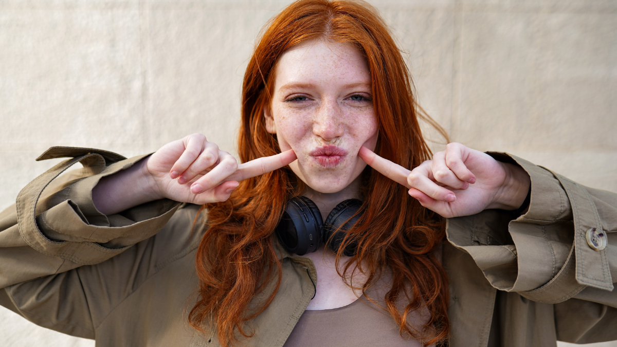 Happy funny teen stylish cool redhead girl standing on urban wall background looking at camera. Portrait of pretty smiling teenage girl with red hair and freckles having fun laughing outdoor.