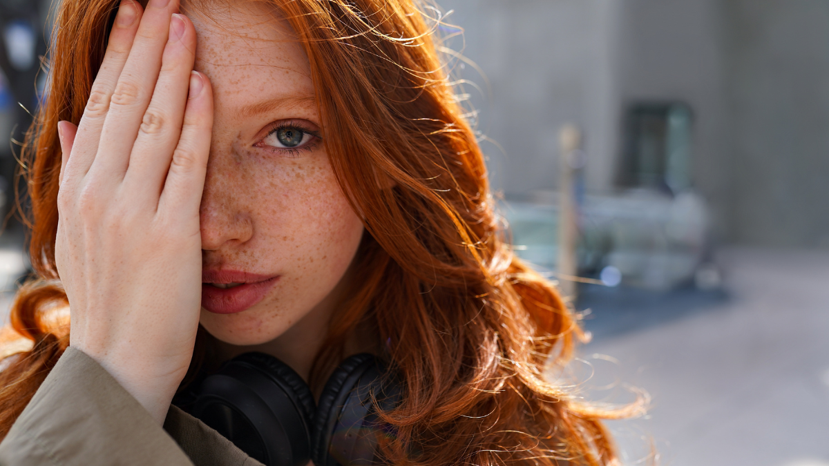 Hipster teen redhead fashion girl model posing in big city urban location. Beautiful teenage girl with red hair looking at camera covering half of face with hand. Close up portrait.