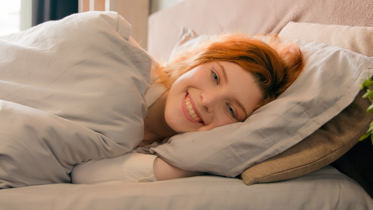 Sleepy beautiful woman waking up lying in comfortable white bed smiling toothy happy smile carefree attractive girl female early good morning relax in cozy bedroom lazy lady awake after sleep at home.