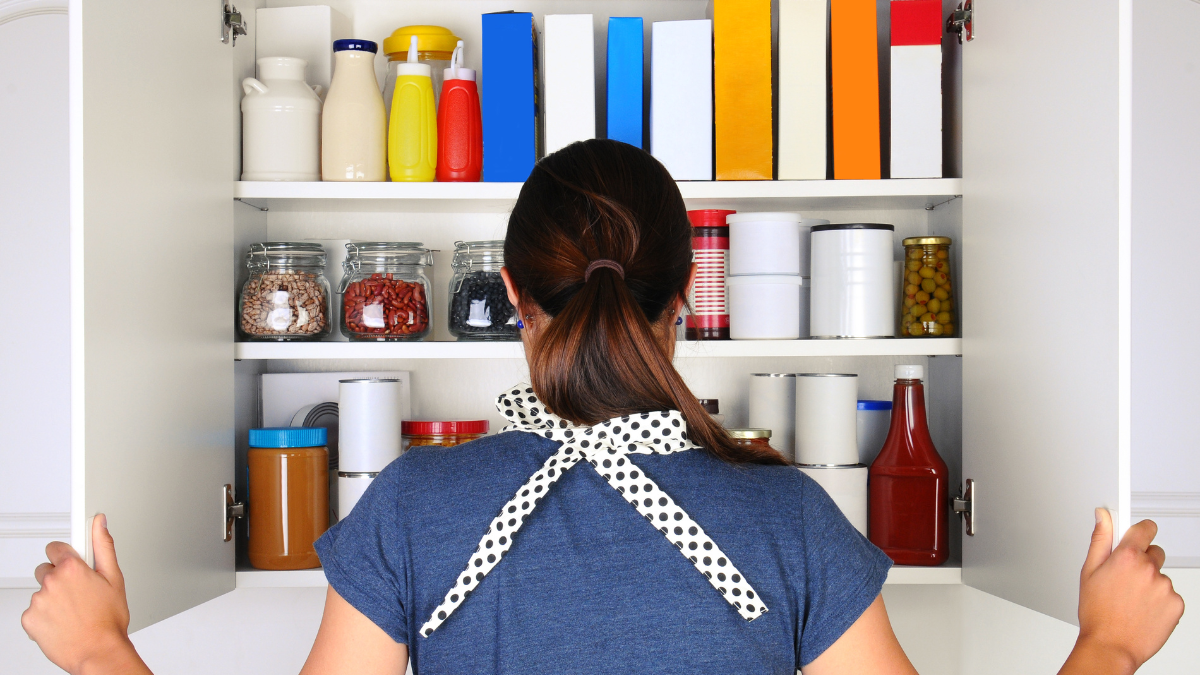 A woman seen from behind opening the doors to a fully stocked pantry. The cupboard is filled with various food stuff and groceries all with blank labels. Horizontal format the woman is unrecognizable.