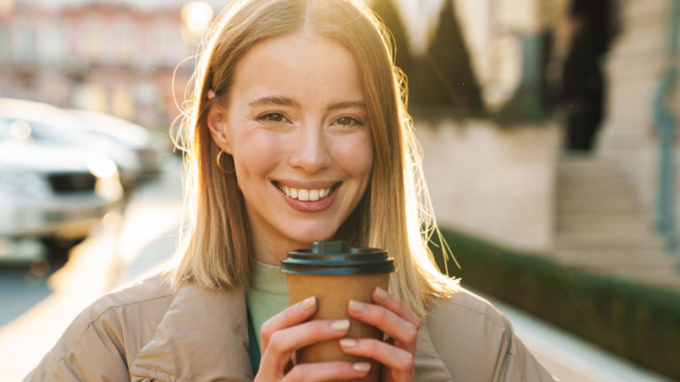 Portrait of joyful caucasian woman smiling and drinking coffee takeaway while walking at city street.